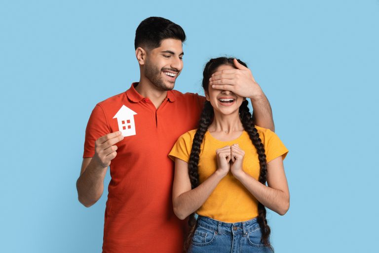 Couple smiling with main covering woman's eyes with his hand and holding a paper cut out of a home ©Prostock-studio