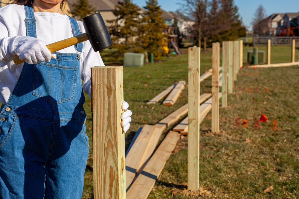 Woman hammering wooden post- fence installation in suburban neighborhood ©grandbrothers