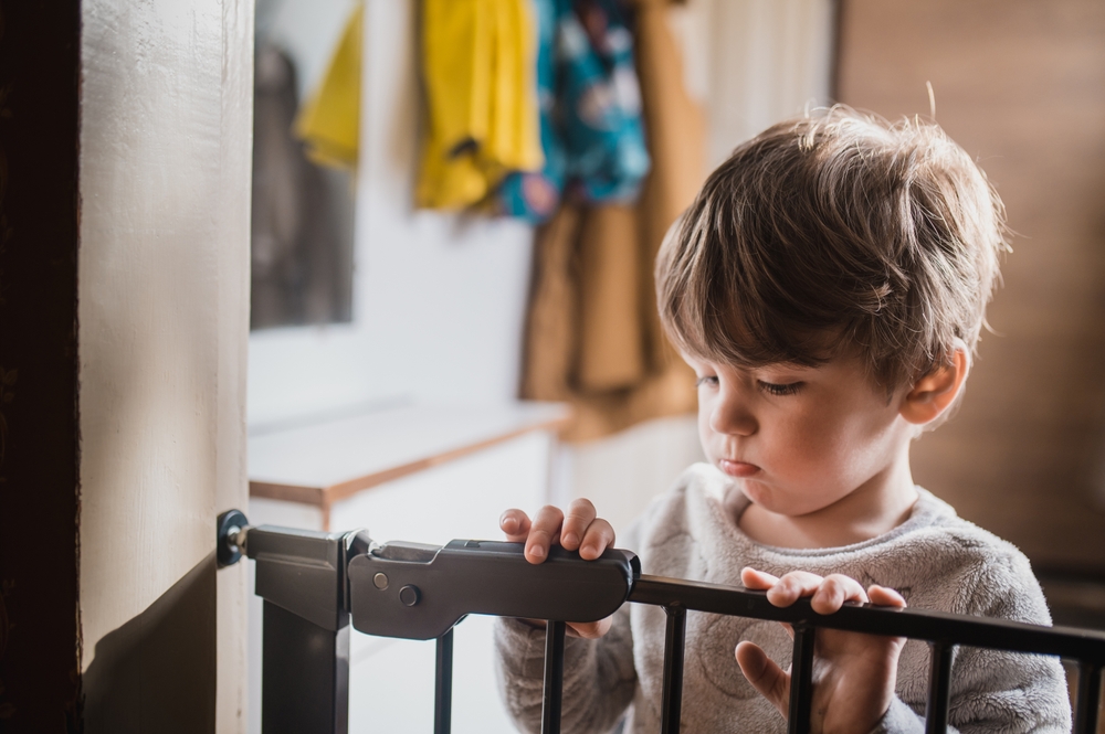 Front view of a little boy standing on the other side of the child safety gate ©illustrissima