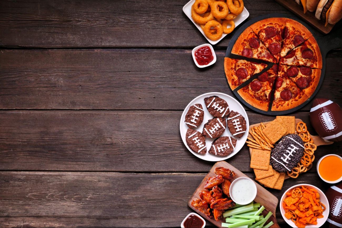 Game day party spread with pizza, wings, brownies, onion rings, pretzels, and snacks on a wooden table. ©JeniFoto