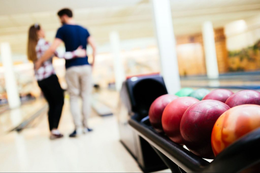 Close up of a rack of bowling balls at a bowling alley with a blurred view of a couple with their arms around each other ©NDAB Creativity