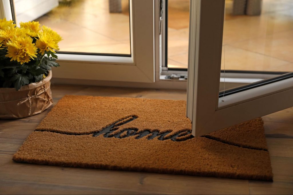 Cozy indoor entryway with door open. There is a “Home” welcome mat written and cursive and yellow potted flowers nearby.
©New Africa