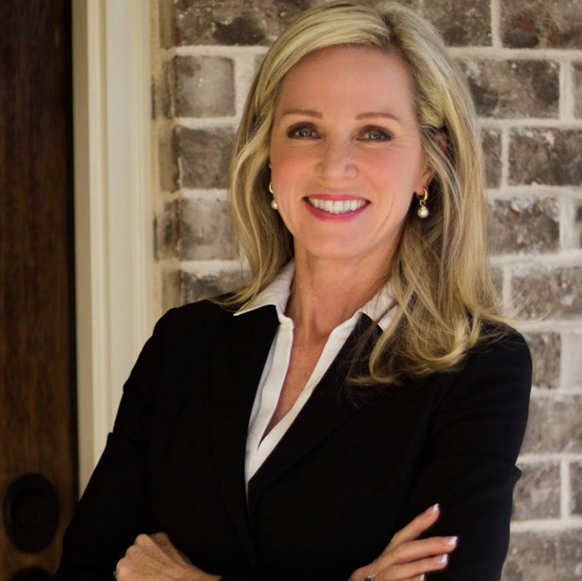 Headshot of Kathryn Cannon, a professional woman with shoulder-length blonde hair, wearing a black blazer and white blouse, smiling with her arms crossed in front of a brick wall.